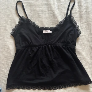 Svart linne från Nelly med spetsdetaljer - Snygg svart linne från Nelly med spetsdetaljer längs kanterna och en liten rosett framtill. Perfekt för en stilren look.