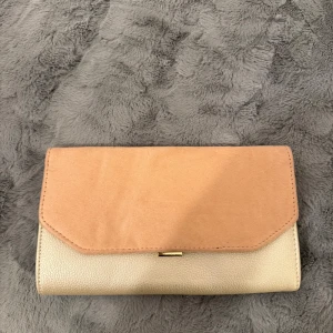 Beige och rosa kuvertväska - Snygg kuvertväska i beige och rosa med en stilren design. Väskan har en praktisk storlek och en liten gulddetalj framtill. Perfekt för att bära med sig det viktigaste.