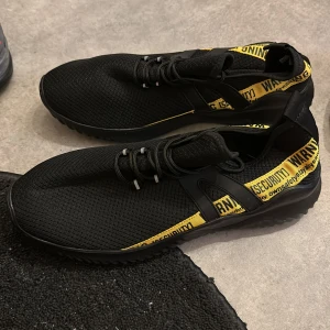 Svarta sneakers med gul detalj - Snygga svarta sneakers med en iögonfallande gul rem med texten 'WARNING SECURITY'. Skorna har en stickad ovandel och snörning för en bekväm passform. Perfekta för en trendig look.