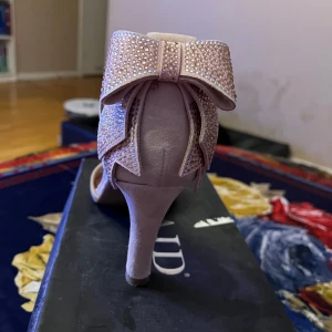 Beige klackskor med rosett - Eleganta beige klackskor med en stor glittrig rosett på baksidan. Skorna har en öppen tå och en hög klack, vilket ger en stilren och festlig look.