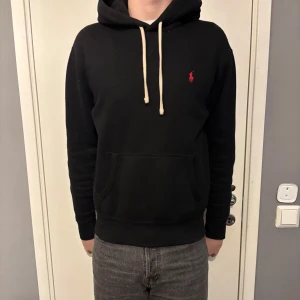 Svart hoodie från Polo Ralph Lauren - Säljer en klassisk svart hoodie från Polo Ralph Lauren med det ikoniska röda logotypen på bröstet. Tröjan har en känguruficka och justerbar huva med vita snören. Perfekt för en avslappnad stil. Säljer pågrund av att den har blivit för liten för mig. Kom gärna med prisförslag Mvh Elias🙌🏼