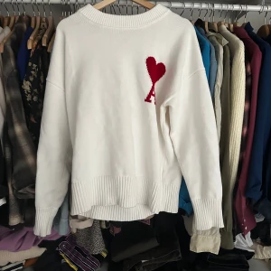 Vit stickad tröja från Ami Paris - Säljer en stilren vit stickad tröja från Ami Paris med ett rött hjärtmönster på bröstet. Tröjan har långa ärmar och ribbade kanter vid ärmslut och nederkant. Perfekt för en trendig och bekväm look.