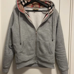 Grå huvtröja med dragkedja Bruberry  - Säljer en stilren grå huvtröja från Burberry med dragkedja och klassiskt rutigt mönster i huvan. Tröjan har långa ärmar och praktiska fickor framtill. Perfekt för en avslappnad look.