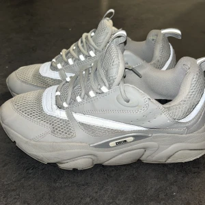 Grå sneakers från Dior - Snygga grå sneakers från Dior med vit detalj och snörning. Skorna har en sportig design med mesh och syntetmaterial för en modern look. Perfekta för en stilren vardagsoutfit.