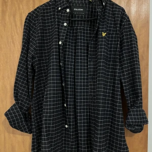 Svart rutig skjorta från Lyle & Scott - Snygg svart skjorta från Lyle & Scott med vitt rutmönster och ikonisk logga på bröstet. Skjortan har långa ärmar och knappar framtill. Perfekt för en stilren look.