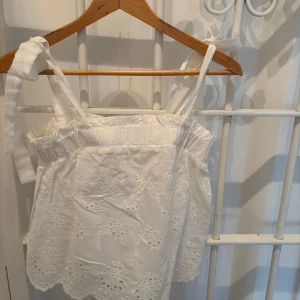 Vit axelbandslös blus - Charmig vit blus med broderade detaljer och knytband på axlarna. Den har en luftig och somrig design med spetsdetaljer längs nederkanten. 