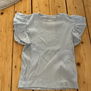 Ljusblå ribbad topp med volangärmar - Säljer en söt ljusblå ribbad topp med volangärmar. Perfekt för en avslappnad och stilren look. Toppen har en rund halsringning och är tillverkad i ett mjukt material.