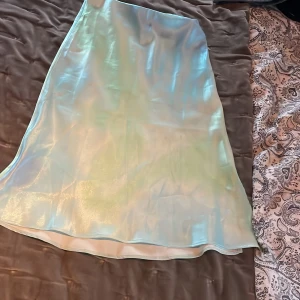 Iridescent kjol från H&M. Siden kjol - Säljer en glansig iridescent kjol från H&M i en vacker ljusblå nyans. Kjolen har en elegant och flytande design som fångar ljuset på ett fantastiskt sätt. Perfekt för att lägga till lite extra stil till din garderob.