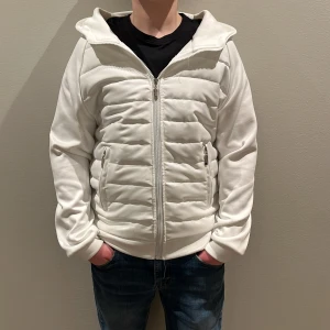 Vit quiltad jacka med dragkedja - Snygg vit jacka med quiltad design och dragkedja framtill. Jackan har en huva och långa ärmar, perfekt för kyligare dagar. Den har också praktiska fickor med dragkedja. 