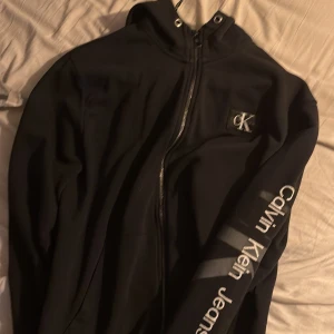 Svart hoodie från Calvin Klein Jeans - Säljer en svart hoodie från Calvin Klein Jeans med dragkedja och logga på bröstet. Ärmarna har texten 'Calvin Klein Jeans' tryckt längs sidan. Perfekt för en stilren och bekväm look. Den har storlek xl men sitter precis som en L möjligtvis en större M .