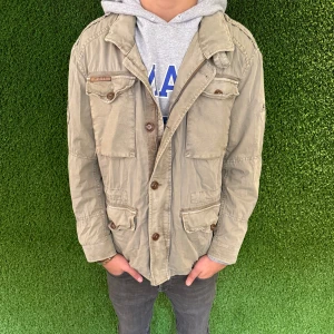 Fieldjacka  - Säljer en beige jacka med flera fickor och knappar framtill. Jackan har en avslappnad stil och är perfekt för höst och vår. Den har långa ärmar och en klassisk krage.