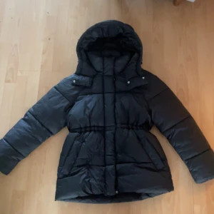 Svart dunjacka med huva - Säljer en svart dunjacka med huva. Jackan har långa ärmar och en dragkedja framtill. Perfekt för kalla dagar. Två mindre hål, se sista bilden. De är igen sydda! 