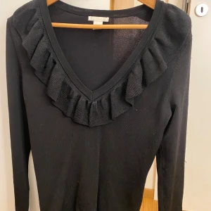 Svart tröja med volangdetalj - Snygg svart tröja med volangdetalj runt v-ringningen. Perfekt för en stilren look. Långärmad och i ett mjukt material som ger en bekväm passform.