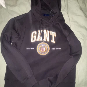 Svart hoodie från GANT - Säljer en svart hoodie från GANT med tryck framtill. Den har en klassisk design med justerbar huva och ribbade muddar. Perfekt för en avslappnad stil.