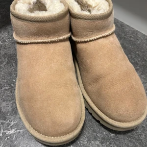 Beige boots från UGG - Säljer ett par mysiga beige boots från UGG med mjukt foder inuti. Perfekta för kalla dagar med sin varma insida och stilrena design. De har en rund tå och är tillverkade i mocka. Nästan inga synliga täcken på användning. Låga Ugg. 