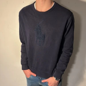 Marinblå tröja från Ralph Lauren - Säljer en stilren marinblå tröja från Ralph Lauren med en riktigt fet broderad logga på bröstet | Tröjan är i ett varmt bekvämt material och en klassisk rund halsringning | Perfekt för en avslappnad och snygg look | Hör av er vid minsta lilla fråga eller fundering!