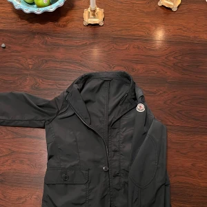 Svart jacka från Moncler - Snygg svart jacka från Moncler med dragkedja och knappar. Den har en klassisk design med Moncler-logga på ärmen och en praktisk ficka framtill. Perfekt för en stilren look. En av knapparna har lossnat men det märks inte särskilt tydligt för fler bilder skriv till mig privat. Jackan är en äldre modell storlek 152 cm 