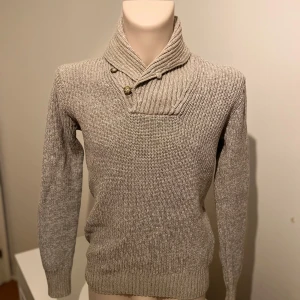 Grå stickad tröja från H&M - Säljer en stilren grå stickad tröja från H&M med en unik krage och knapplösning. Perfekt för kyliga dagar med sin mjuka och bekväma passform. Tröjan har långa ärmar och en ribbad struktur som ger en klassisk look.