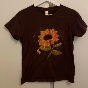 Brun T-shirt - OANVÄND - Söt brun t-shirt med ett motiv av en solros. T-shirten har korta ärmar och en rund halsringning. Perfekt för en avslappnad look. Helt oanvänd!