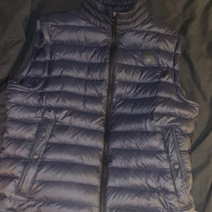 Blå dunväst från Hugo Boss - Snygg blå dunväst från Hugo Boss med dragkedja och hög krage. Perfekt för kyliga dagar och ger en stilren look. Västen har två fickor med knappar framtill.