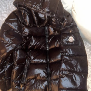 Moncler maya - Snygg svart pufferjacka från Moncler med glansig finish och huva. Jackan har en dragkedja framtill och en broderad logotyp på ärmen. Perfekt för kyliga dagar. Har haft den ungefär 1 å ett halvt år då tänker köpa något mer som e passbart