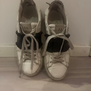 Valentino sneakers med svarta detaljer - Snygga vita sneakers från Valentino med svarta detaljer och snörning. Skorna har en unik design med rockstuds på hälen och en bred svart rem över foten. Perfekta för en stilren look. Väll använda 