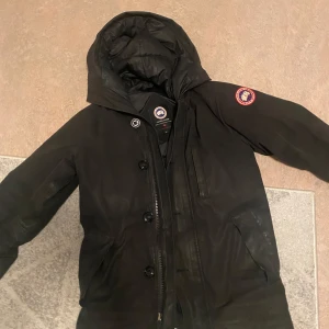 Svart dunjacka från Canada Goose - Säljer en svart dunjacka från Canada Goose med huva och dragkedja. Jackan har en broderad logotyp på ärmen och knappar framtill. Perfekt för kalla dagar. Det är en sällsynt modell priset kan diskuteras kill modell men funkar för tjejer