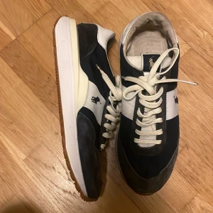 Svart och vit Ralph Lauren skor - Säljer ett par snygga sneakers från Ralph Lauren i svart och vitt. De har en klassisk design med snörning och en bekväm sula. Perfekta för vardagsbruk och ger en stilren look. Skorna har en ikonisk logga på sidan och är i okej skick. Går att tvätta för att få det se bättre
