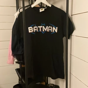 Svart Batman t-shirt - Säljer en svart t-shirt med Batman-logga i vitt och blått. T-shirten har korta ärmar och är tillverkad i bomull. Perfekt för alla Batman-fans!