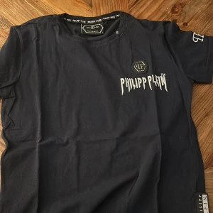 Svart t-shirt från Philipp Plein - Snygg svart t-shirt från Philipp Plein med vit logotyp på bröstet och ärmen. T-shirten har en rund halsringning och är kortärmad. Perfekt för en stilren look.
