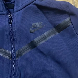 Mörkblå nike tech fleece - Säljer en mörkblå huvtröja från Nike med dragkedja framtill. Tröjan har långa ärmar och en stilren design med diskret logga på bröstet. Perfekt för en avslappnad stil.