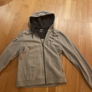 Grå huvtröja från Helly Hansen - Säljer en grå huvtröja från Helly Hansen med dragkedja och broderad detalj på ärmen. Tröjan har en justerbar huva och tryck på bröstet. Perfekt för en avslappnad stil.