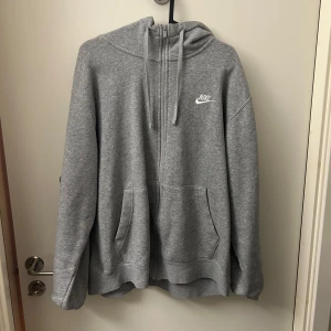 Grå hoodie från Nike - Säljer en klassisk grå hoodie från Nike med dragkedja och huva. Den har en broderad logga på bröstet och praktiska fickor framtill. Perfekt för en avslappnad stil.