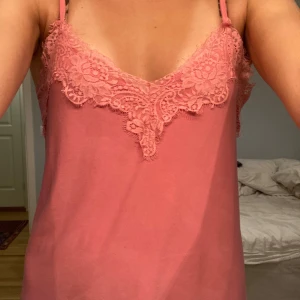 Rosa linne med spetsdetaljer - Säljer ett sött rosa linne med smala axelband och vacker spetsdetalj vid halsringningen. Perfekt för varma dagar eller som ett lager under en kavaj.