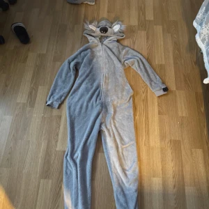 Grå koala onesie från Lindex - Mysig grå onesie från Lindex med koala-design. Den har en huva med koalaansikte och öron, samt dragkedja framtill. Perfekt för en avslappnad dag hemma eller som en rolig kostym.