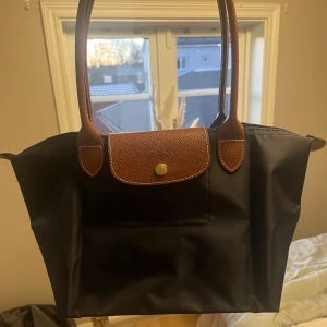 Mörkblå axelväska med bruna detaljer - Snygg mörkblå axelväska med bruna handtag och dragkedja. Väskan har en praktisk design med en knappdetalj på framsidan. Perfekt för att bära med sig det nödvändigaste med stil.