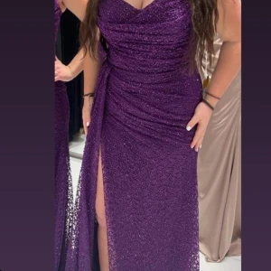 Lila glittrig långklänning med slits - Säljer en superfin lila långklänning som glittrar så snyggt! Den har en draperad överdel och en hög slits som gör den extra festlig. Perfekt för bal eller fest. Klänningen är i nyskick och har en tight passform. Materialet känns lyxigt och bekvämt.