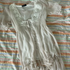 Vit spetsklänning från Yuki - Säljer en superfin vit klänning från Yuki. Den är gjord av ett lätt och luftigt material, perfekt för sommaren. Klänningen har korta ärmar och en vacker spetsdetalj runt halsen och knappar framtill. Den har också en volangkant längst ner som ger en söt touch. Perfekt för sommarens alla fester och tillfällen! 🌸