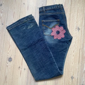 Lågmidjade jeans  - Säljer ett par snygga blå jeans med coola blommiga detaljer på framsidan och baksidan. midjemått: 37 cm, Innerbenslängd: 82 cm