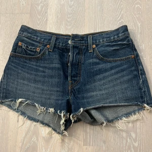 Jeansshorts från Levi's - Säljer ett par snygga jeansshorts från Levi's. De är mörkblå och har en rå kant nertill som ger en cool, avslappnad look. Shortsen har knappar istället för dragkedja och klassiska fem fickor. Perfekta för sommaren!