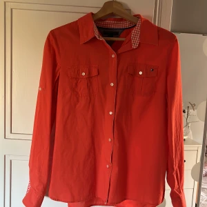 Röd långärmad skjorta från Tommy Hilfiger - Säljer en snygg röd långärmad skjorta från Tommy Hilfiger. Skjortan har en klassisk krage med ett rutigt mönster på insidan och knappar framtill. Den har två bröstfickor med knappar och är i nyskick. Perfekt för både vardag och fest!