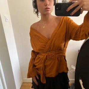 Orange omlottopp med bälte - Säljer en snygg orange/brun omlottopp med bälte i midjan. Toppen har en v-ringad design och långa ärmar. Perfekt för både vardag och fest! 🧡