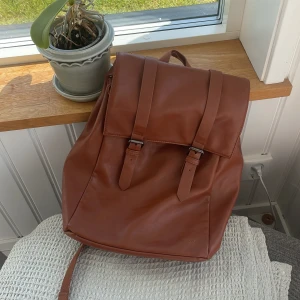 Brun ryggsäck i skinn - Säljer en snygg och praktisk brun ryggsäck i skinn. Väskan har två spännen framtill och ett randigt innerfoder. Perfekt för både vardag och utflykter. Väskan är i bra skick och har gott om plats för dina saker. Köp direkt eller ge prisförslag!💓