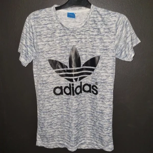 Gråmelerad t-shirt från Adidas - Säljer en snygg gråmelerad t-shirt från Adidas i storlek M. T-shirten har en stor svart Adidas-logga på framsidan och är tillverkad i mjukt och bekvämt material. Perfekt för både träning och vardag! 💪