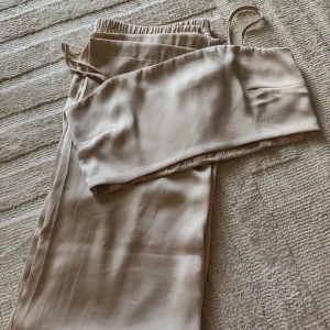 Beige satin set - Säljer ett superfint biege set i satin. Toppen har smala justerbara axelband samt resår i ryggen. Byxorna är lite tightare upptill och lösare nertill, blir ett väldigt fint fall, samt resår i midjan. Matrialet är väldigt mjukt och har en glansig yta som känns lyxigt. Perfekt för fest eller en utekväll!🍾 Detta set är i nyskick och är i storlek M/L. Hör av er vid frågor eller funderingar💕