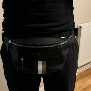 Svart midjeväska med dragkedja - Säljer en snygg svart midjeväska i skinn. Väskan har en dragkedja på framsidan och en justerbar rem med ett spänne. Perfekt för att ha med sig det viktigaste när man är ute och rör på sig. Den har också en liten ficka med dragkedja på framsidan för extra förvaring.