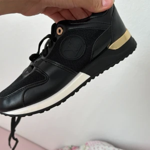 Svarta sneakers med gulddetaljer - Säljer ett par snygga svarta sneakers med gulddetaljer. Skorna har en vit sula och är gjorda av syntetmaterial med meshpartier för extra andningsförmåga. De har snörning framtill och en bekväm passform. Perfekta för både vardag och träning!
