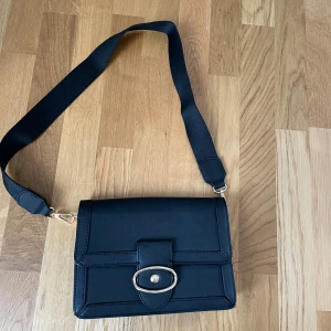 Svart väska - Säljer denna rymliga väska som aldrig har varit använd. Väskan stängs med en magnet. Väskan är köpt för flera år sedan utomlands därav är det oklart vilket märke det är. Väskan har en inbyggd plånbok med plats för flera kort och ficka med dragkedja