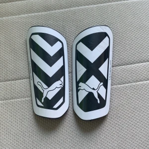 Svartvita benskydd från Puma - Säljer ett par snygga och stilrena benskydd från Puma. De är svartvita med ett coolt mönster och Puma-loggan på framsidan. Baksidan har en bekväm vaddering för extra skydd och komfort. Perfekta för fotbollsträningen eller matchen!