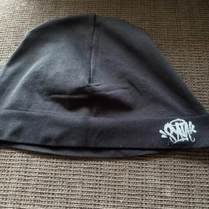 Svart mössa från SYNA - Säljer en svart mössa från SYNA. Den är enkel och stilren med en liten broderad logga i vitt på kanten. Perfekt för att hålla värmen under kyliga dagar. Materialet är en blandning av polyester och elastan, vilket gör den både mjuk och stretchig.